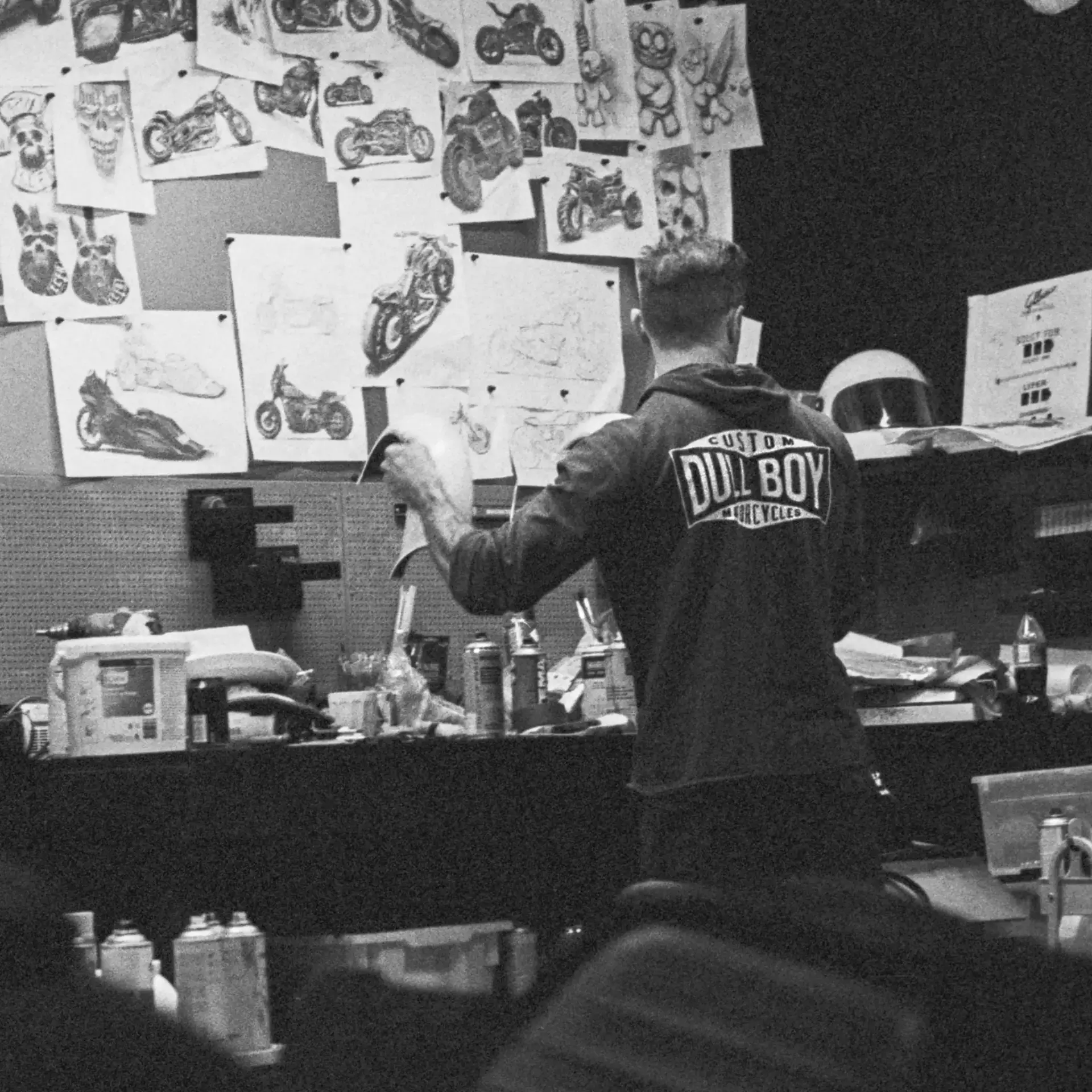 David Hook in a Dull Boy Customs hoodie, seen from behind pinning hand-drawn motorcycle sketches to his workshop wall